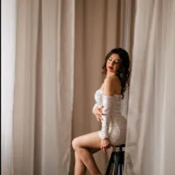 Kobieta w białej, błyszczącej sukience siedzi na czarnym stołku barowym w studio w Piotrkowie Trybunalskim, z beżowymi zasłonami w tle. Modelka ma kręcone włosy i czerwoną szminkę, pozuje...