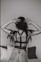 Kobieta w czarno-białym ujęciu od tyłu, ubrana w jeansy i czarny stanik z ozdobnymi piórami na ramionach, dłońmi dotyka włosów, w tle widoczna pościel.