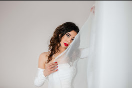 Portret kobiety w białej sukni z odkrytymi ramionami, z czerwonymi ustami i kręconymi włosami, częściowo zasłoniętej przez białą, prześwitującą tkaninę, opierającej się o białą ścianę.