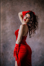 Kobieta w czerwonej, dopasowanej sukience i rękawiczkach pozuje na tle abstrakcyjnego, szaro-brązowego tła, z ręką uniesioną do włosów i spojrzeniem skierowanym w bok.