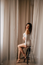 Kobieta w białej, błyszczącej sukience siedzi na czarnym stołku barowym w studio w Piotrkowie Trybunalskim, z beżowymi zasłonami w tle. Modelka ma kręcone włosy i czerwoną szminkę, pozuje...