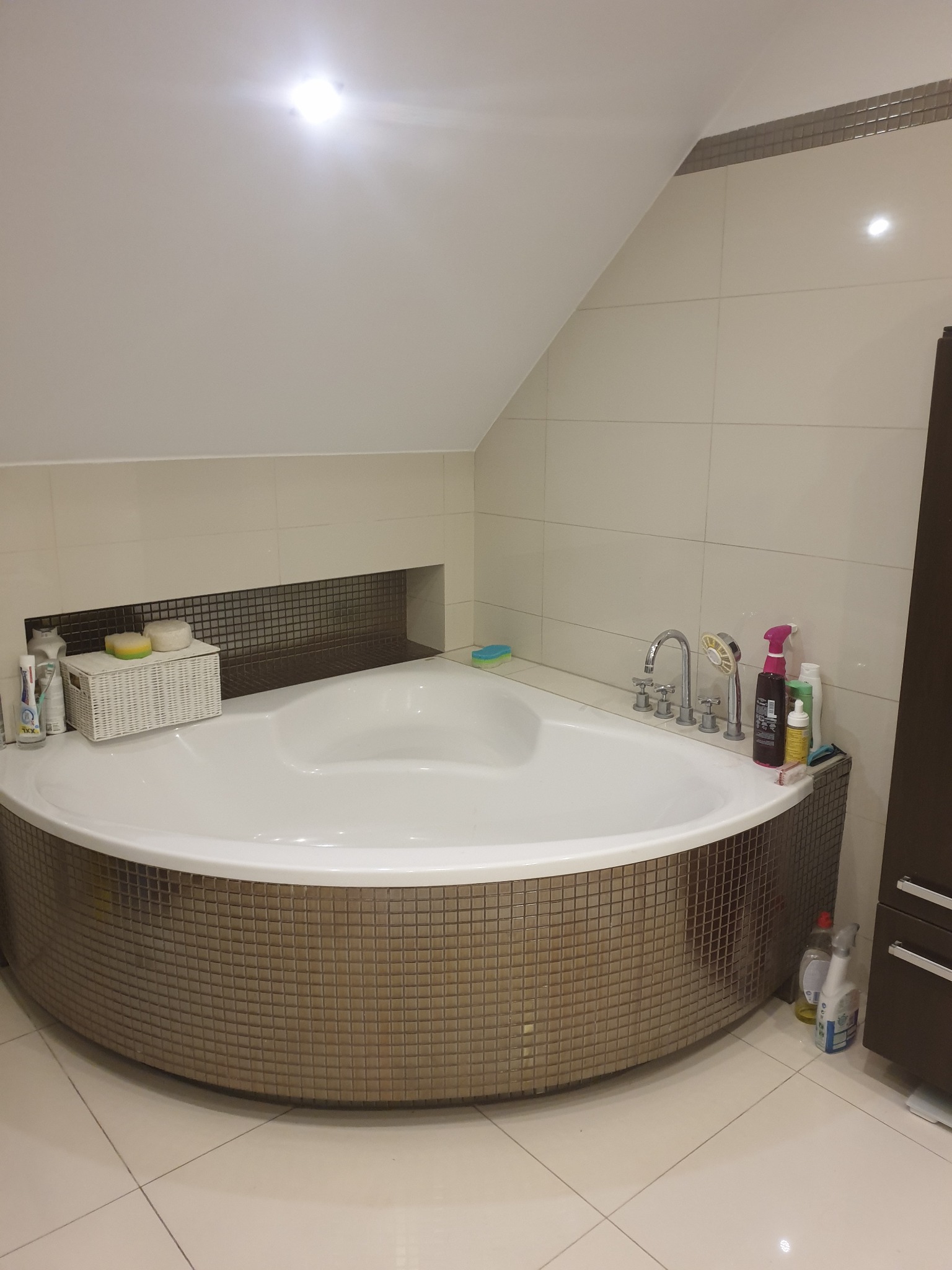 Narożna wanna obudowana mozaiką w łazience na poddaszu w Zabrzu, z widoczną armaturą i akcesoriami kosmetycznymi.