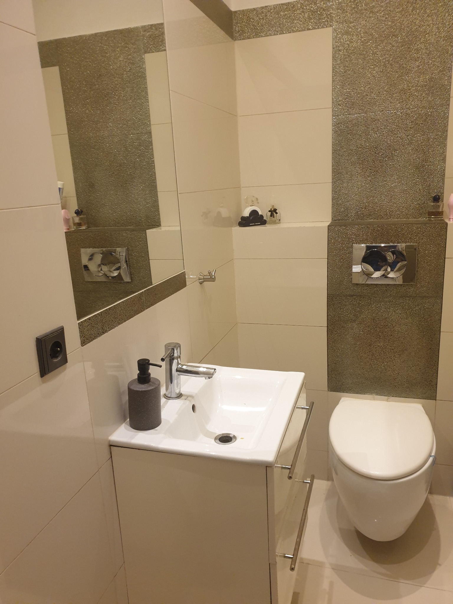 Nowoczesna łazienka z wiszącą toaletą, umywalką nablatową na szafce z chromowanymi uchwytami, lustrem z podświetleniem i dekoracyjnymi panelami ściennymi w odcieniach beżu i brązu z efektem brokatu