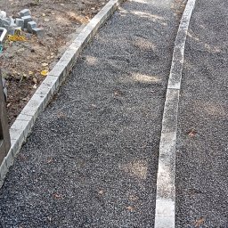 Robbruk - W trakcie tworzenia ścieżki z drobnego kruszywa ograniczonej obrzeżami z jasnoszarej kostki. W tle widoczny budynek z cegły oraz narzędzia budowlane.