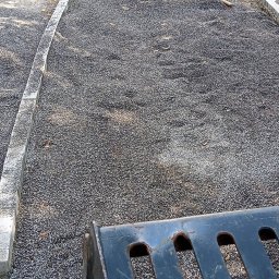 Robbruk - Przygotowanie podłoża z grysu na ścieżkę z obrzeżami z kostki granitowej, widoczny fragment łyżki koparki, ubijarka i wąż do zagęszczarki w tle.