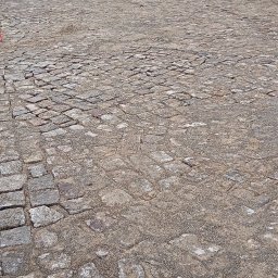 Robbruk - Niedokończona nawierzchnia z kostki granitowej na tle ceglanego budynku z widocznymi pracownikami i sprzętem budowlanym w oddali.