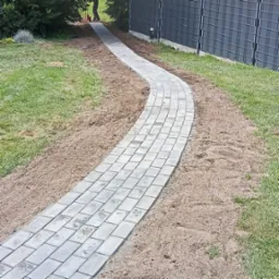 Zakrzywiony chodnik z szarej kostki betonowej, świeżo ułożony w ogrodzie, widoczne ślady piasku i trawy, metalowe ogrodzenie w tle.