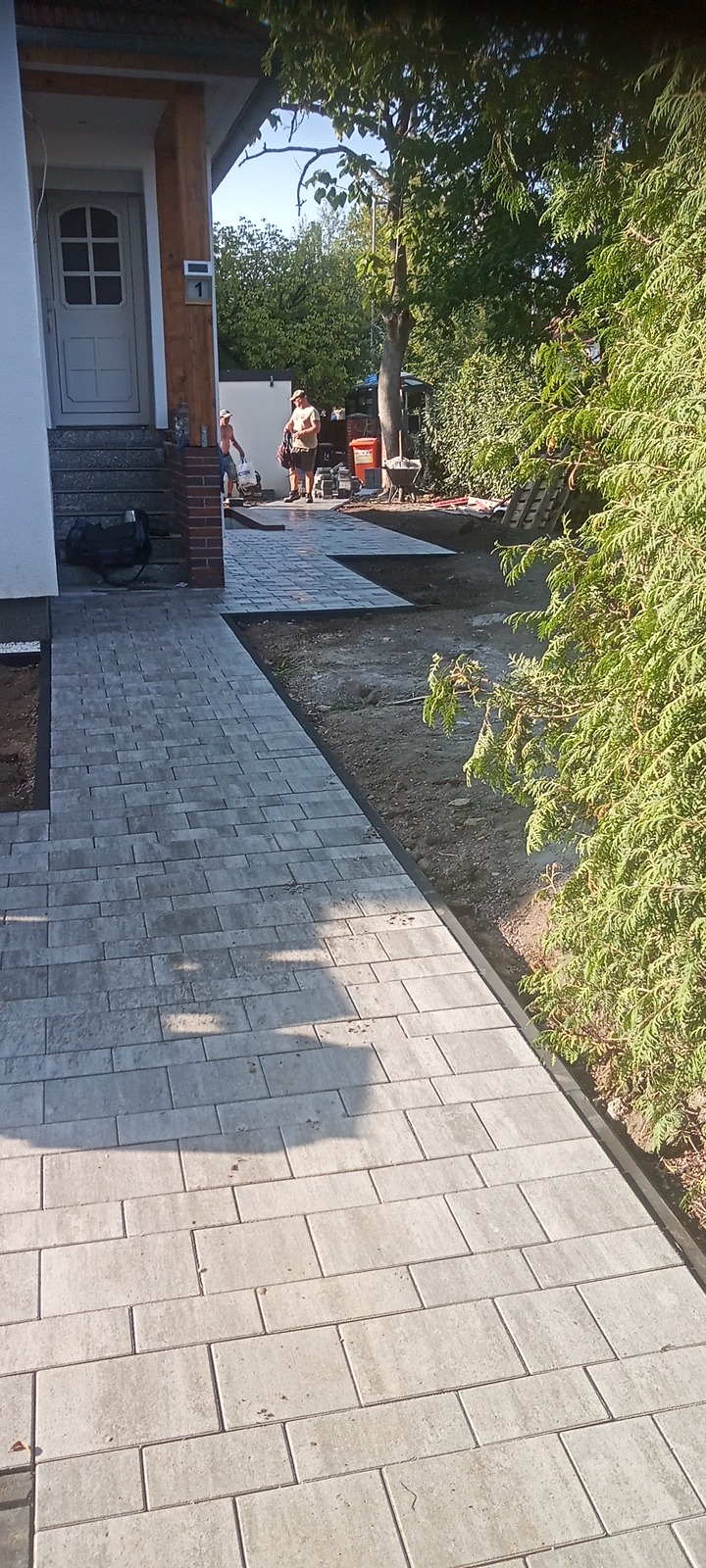 Ułożona ścieżka z szarej kostki brukowej prowadząca do domu z widocznymi pracownikami w tle i przygotowanym terenem pod dalsze prace.