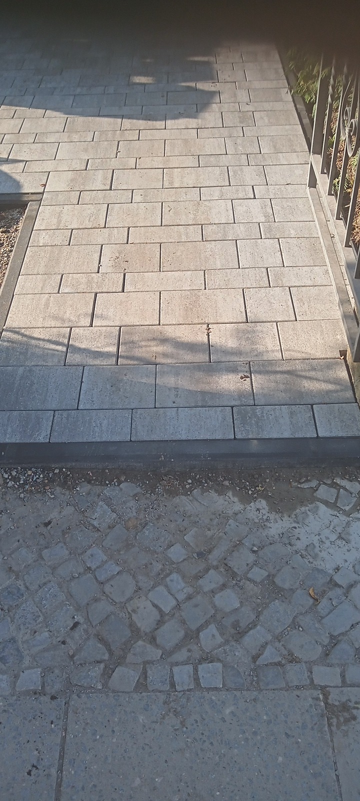 Ułożona ścieżka z prostokątnych, jasnoszarych płyt brukowych, obramowana ciemniejszymi płytami, przechodząca w starą kostkę brukową i betonową nawierzchnię. Żelazne ogrodzenie z prawej strony.