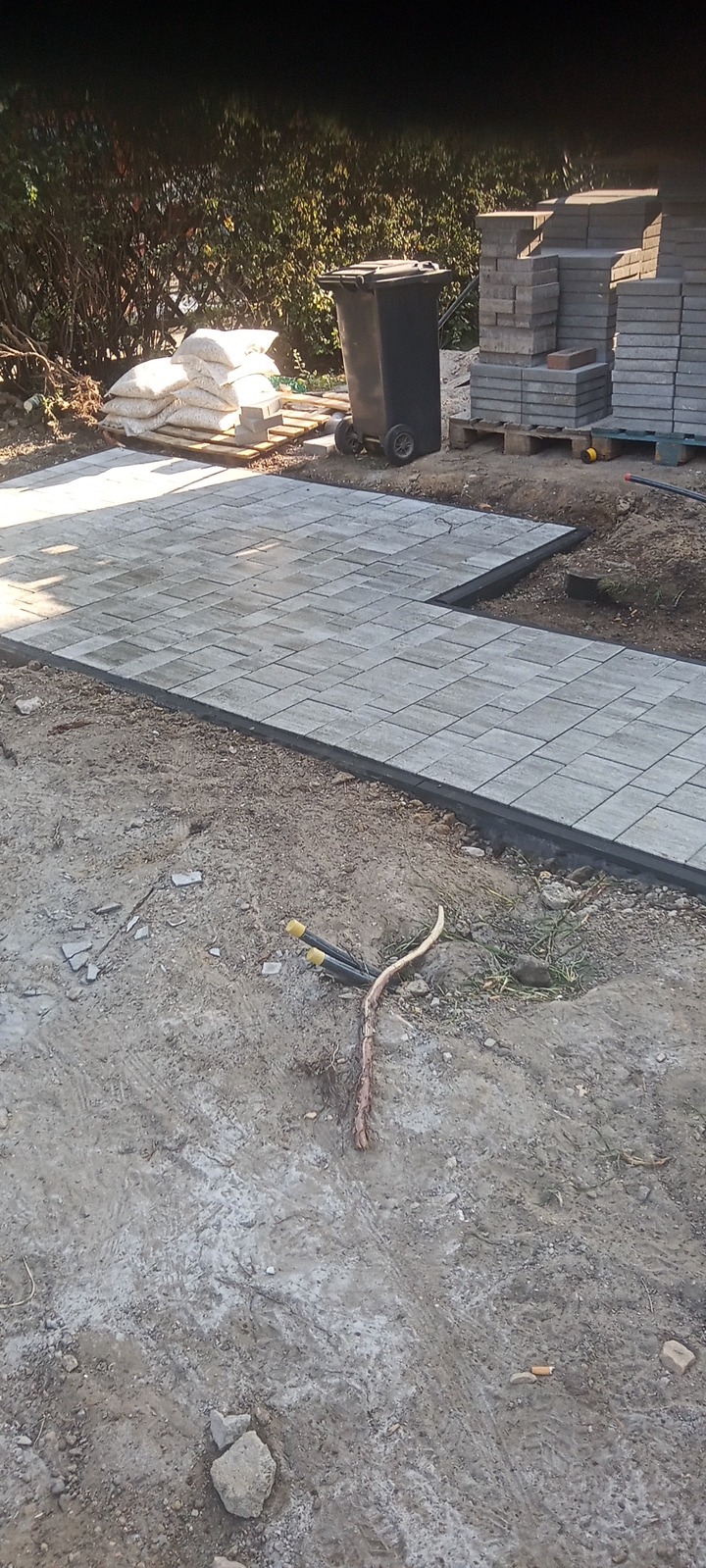 Fragment układanej ścieżki z kostki brukowej w ogrodzie, widoczne worki z materiałem, stos kostki na palecie, czarny kosz na śmieci i wystające rury z żółtymi zaślepkami.