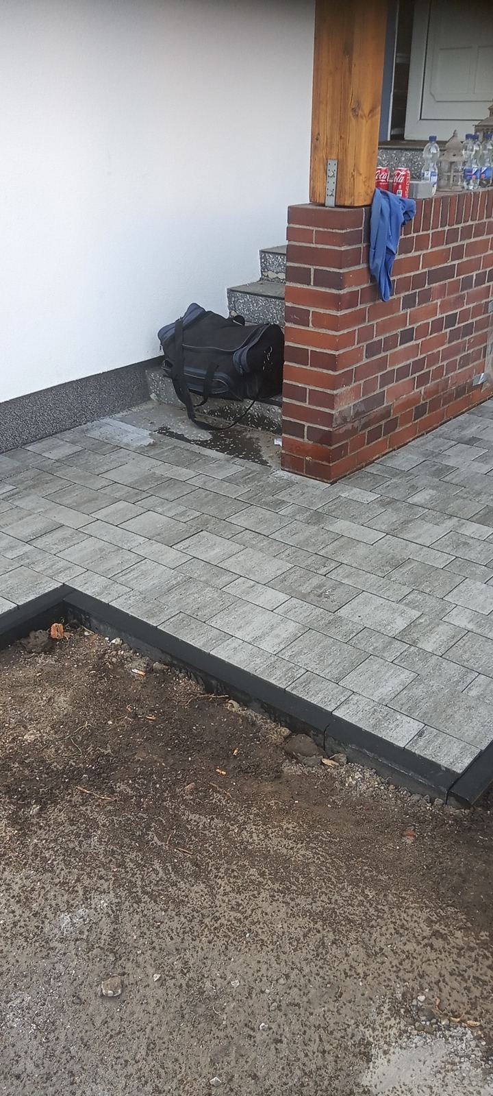 Fragment nowej szarej kostki brukowej z czarnym obrzeżem, widoczny fragment niezakończonej pracy z ziemią, torba robocza i napoje przy wejściu do budynku.