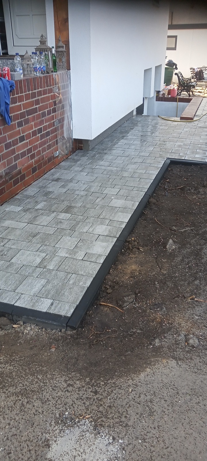 Świeżo ułożona, szara kostka brukowa z czarnym obrzeżem przylegająca do ściany budynku. Część powierzchni wokół kostki jeszcze niezagospodarowana.