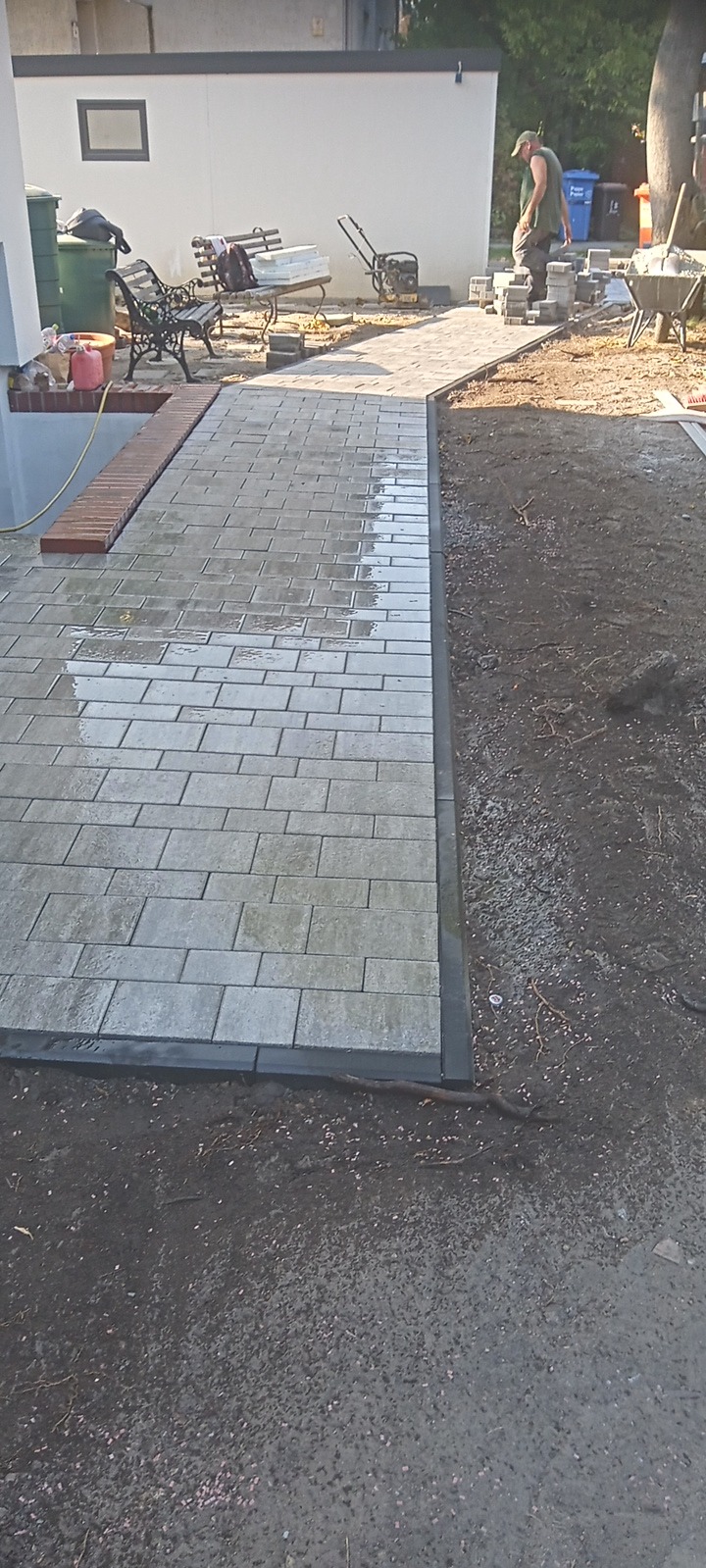 Świeżo ułożona, lekko wilgotna, szara kostka brukowa z ciemnoszarą obwódką, widoczna w trakcie prac wykończeniowych, z pracownikiem w tle i sprzętem budowlanym.