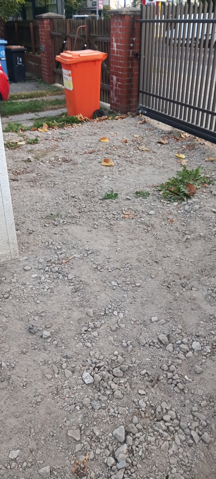 Przygotowany teren pod kostkę: wyrównana, kamienista powierzchnia z widocznymi śladami po pracach ziemnych, obok ogrodzenia i pojemnika na śmieci.