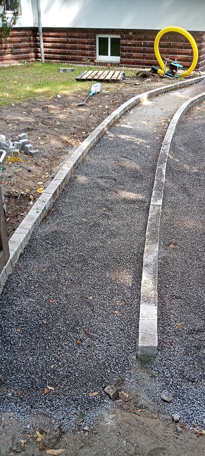 W trakcie tworzenia ścieżki z drobnego kruszywa ograniczonej obrzeżami z jasnoszarej kostki. W tle widoczny budynek z cegły oraz narzędzia budowlane.