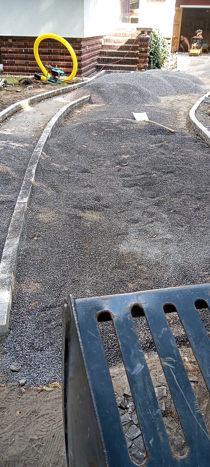 Przygotowanie podłoża z grysu na ścieżkę z obrzeżami z kostki granitowej, widoczny fragment łyżki koparki, ubijarka i wąż do zagęszczarki w tle.