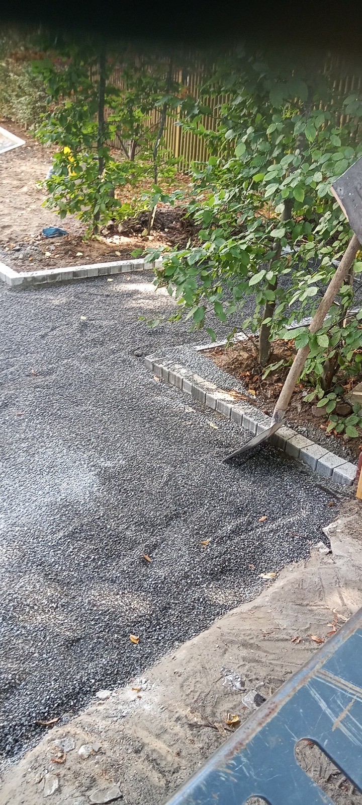 Przygotowanie podłoża pod kostkę: wyrównany żwir ograniczony obrzeżem z kostki, widoczna łopata wbita w żwir, drzewa w tle.