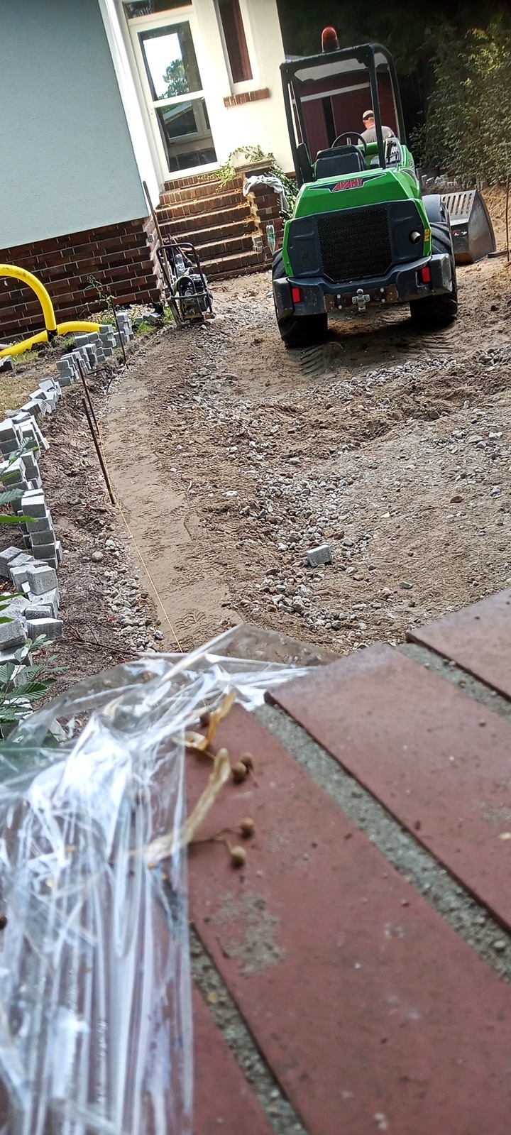 Przygotowanie podłoża pod ścieżkę z kostki: wykop z wytyczoną linią, ubijarka i minikoparka w tle, widok z góry na folię ochronną.