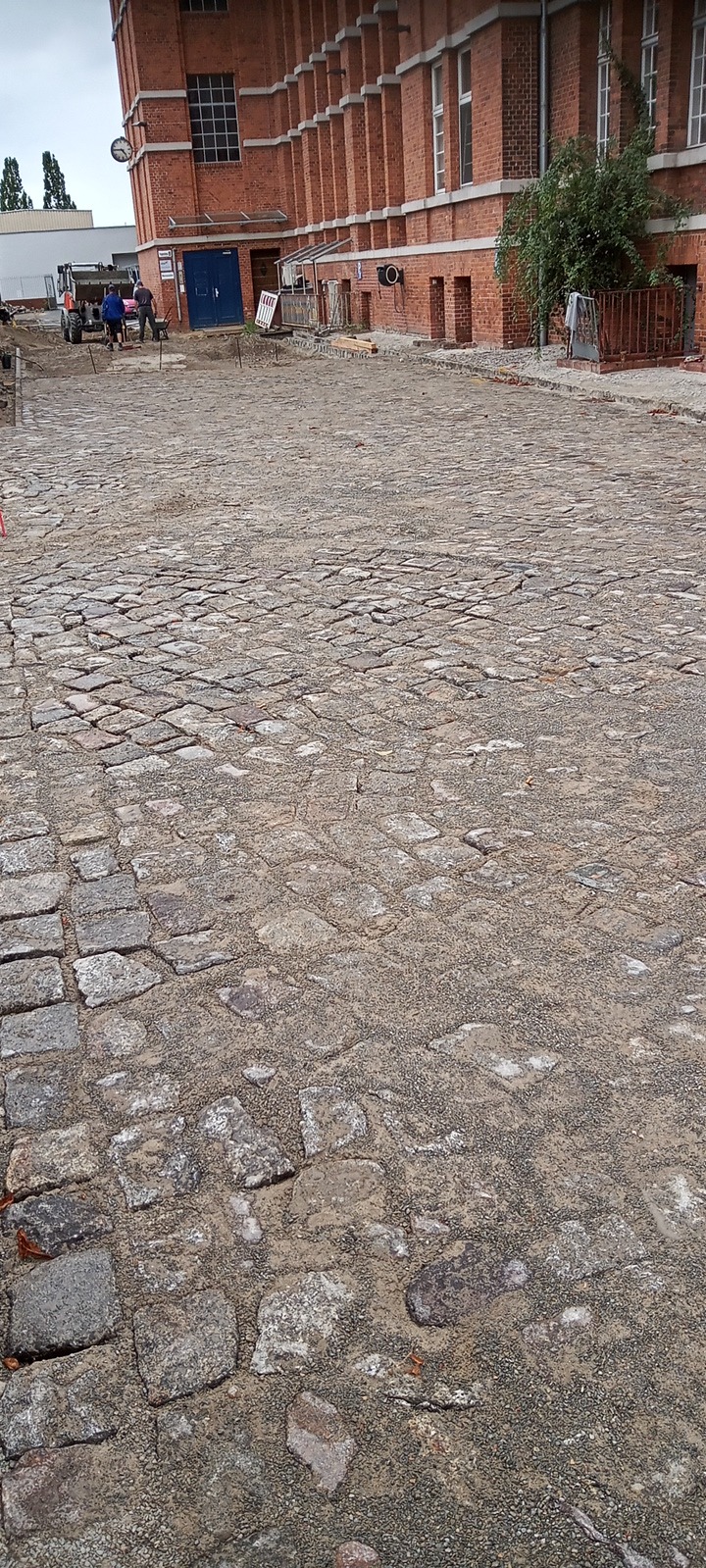 Niedokończona nawierzchnia z kostki granitowej na tle ceglanego budynku z widocznymi pracownikami i sprzętem budowlanym w oddali.