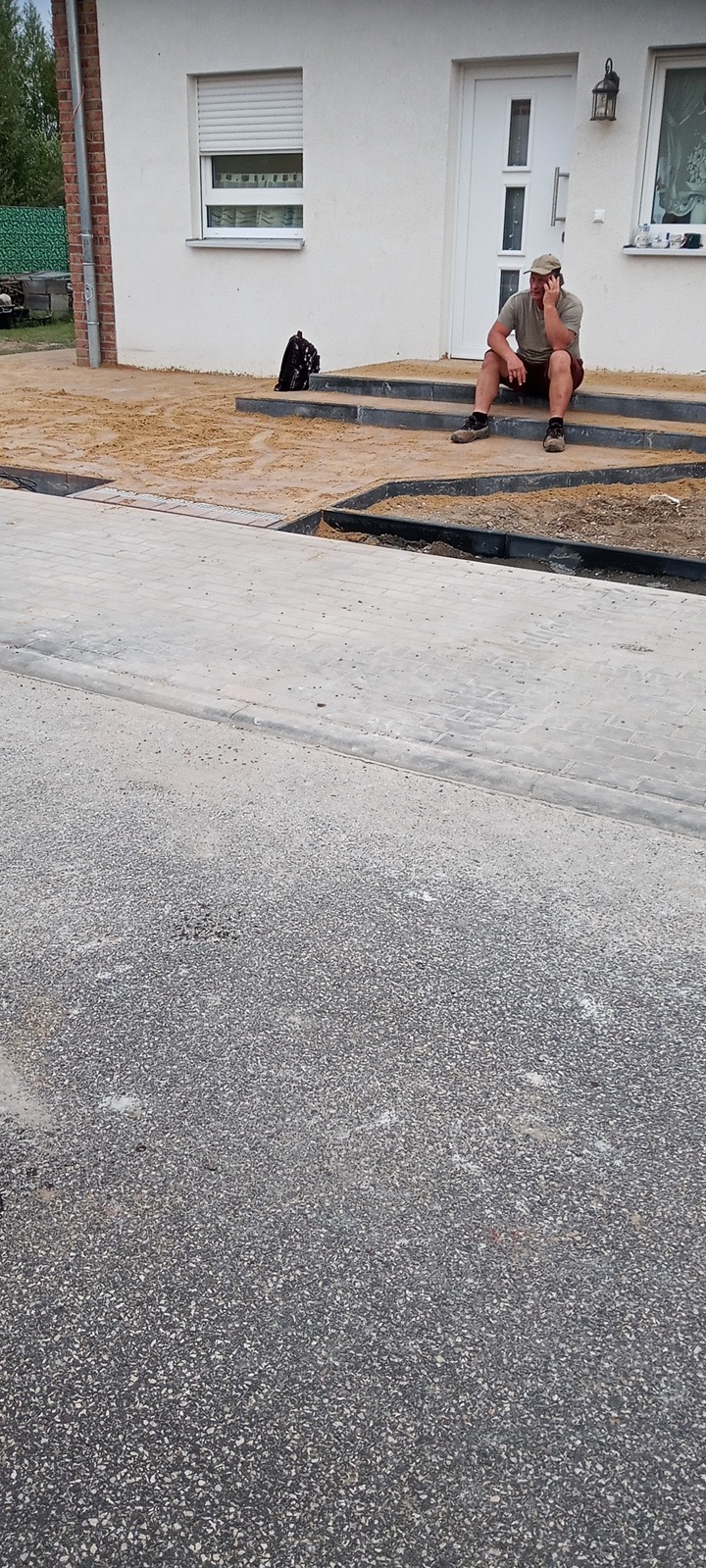 Pracownik siedzi na schodach domu podczas układania nowej kostki brukowej, widoczny fragment chodnika z kostki i przygotowane podłoże pod kolejne elementy.