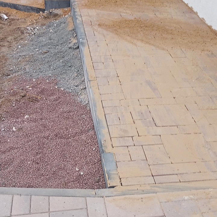 Ułożona ścieżka z kostki brukowej w trakcie zasypywania piaskiem, obok widoczny obszar z czerwonym i szarym żwirem.