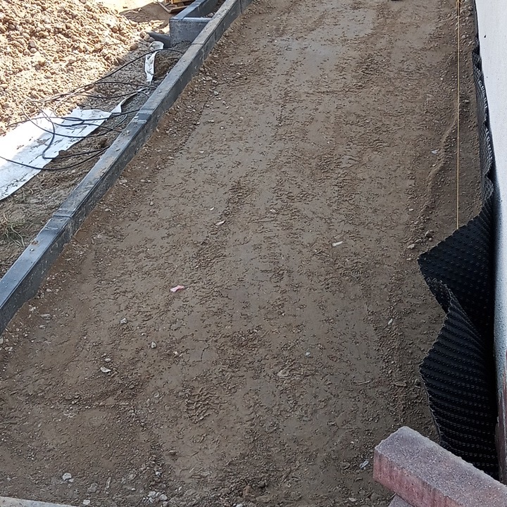 Przygotowanie podłoża pod kostkę brukową w Ciechanowcu: wyrównany teren z obrzeżami i folią drenażową przy ścianie budynku, widoczne materiały budowlane.