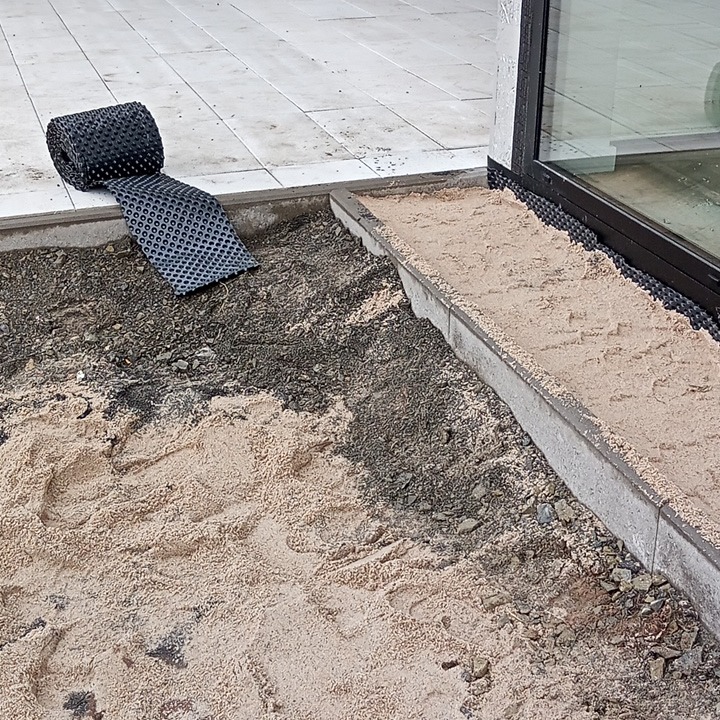 Przygotowanie podłoża pod taras: warstwa piasku i rozwinięta membrana drenażowa przy oknie tarasowym.