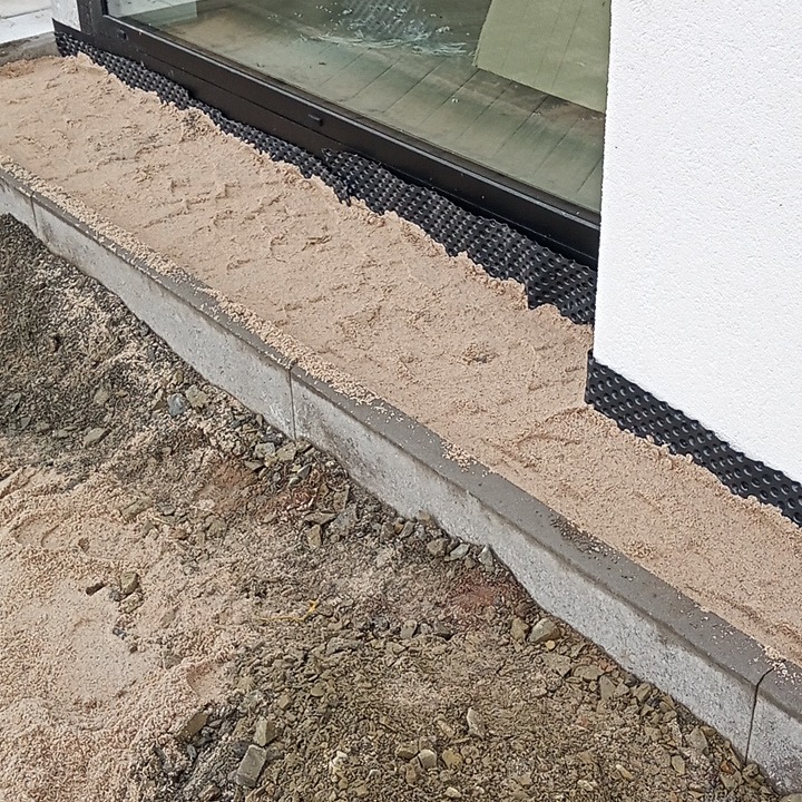 Przygotowanie podłoża pod kostkę: piasek zasypany między obrzeżem a ścianą budynku, widoczna folia kubełkowa.