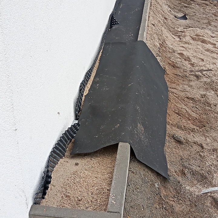 Przygotowanie podłoża pod kostkę: obrzeża betonowe, geowłóknina i drenaż przy ścianie budynku.