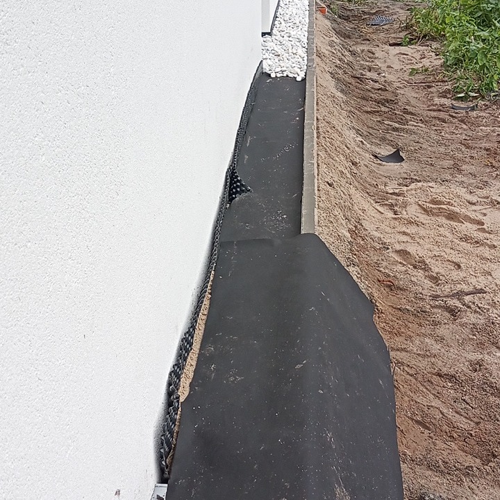 Obrzeże budynku z białym tynkiem, zabezpieczone folią drenażową i czarną geowłókniną, oddzielone od gruntu betonowym krawężnikiem, z widocznym drenażem z białych kamieni.