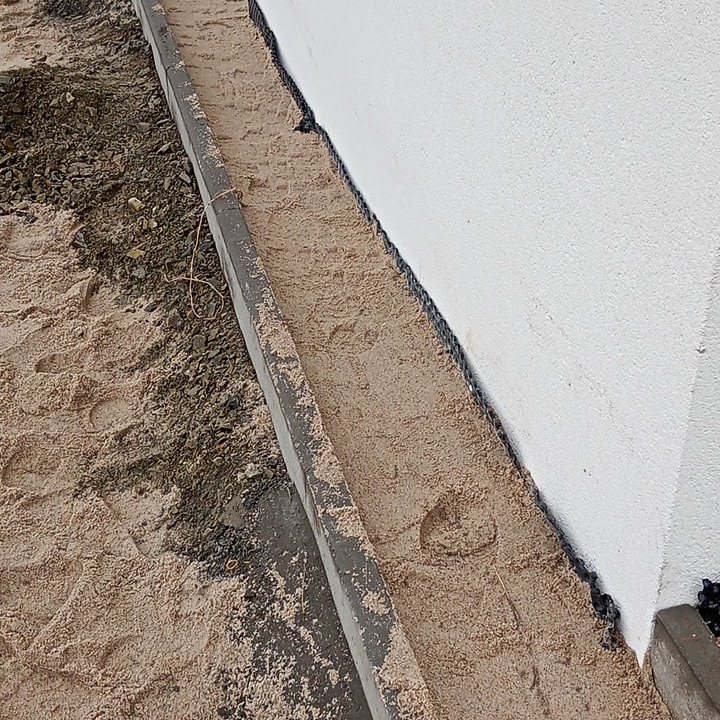 Przygotowane podłoże z podsypką piaskową i obrzeżem betonowym wzdłuż ściany budynku, widoczny odcisk buta w piasku.