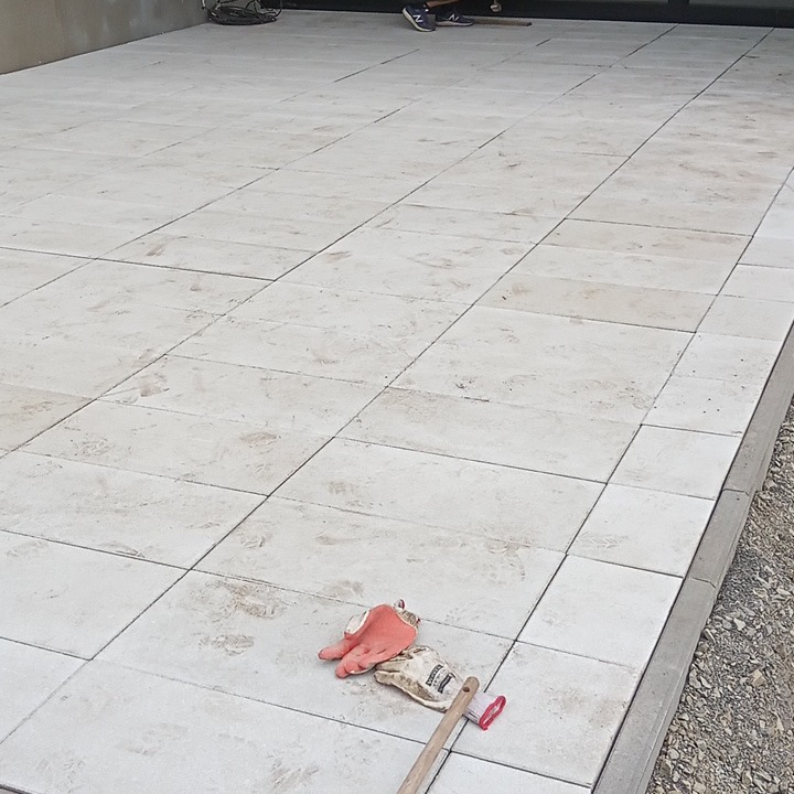 Jasne, prostokątne płyty ułożone na zewnątrz, widoczne robocze rękawice i szczotka do zamiatania, osoba w tle.