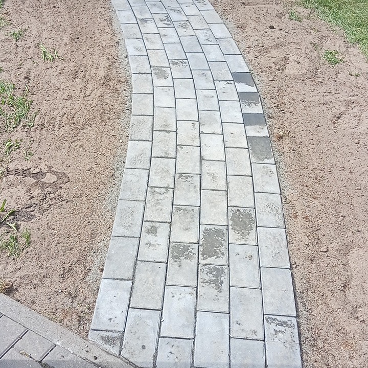 Zakrzywiony chodnik z szarej kostki brukowej, widoczny z góry, otoczony piaskiem i fragmentami trawy.