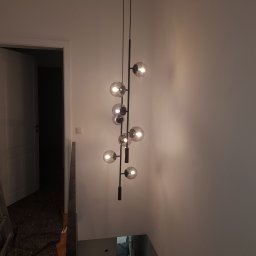 Karol Pielecki - Nowoczesna lampa wisząca z kulistymi kloszami na czarnym stelażu, oświetlająca klatkę schodową w trakcie wykańczania wnętrza, widoczny fragment schodów i otwarte drzwi do pomieszczenia.