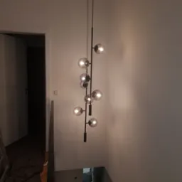 Nowoczesna lampa wisząca z kulistymi kloszami na czarnym stelażu, oświetlająca klatkę schodową w trakcie wykańczania wnętrza, widoczny fragment schodów i otwarte drzwi do pomieszczenia.