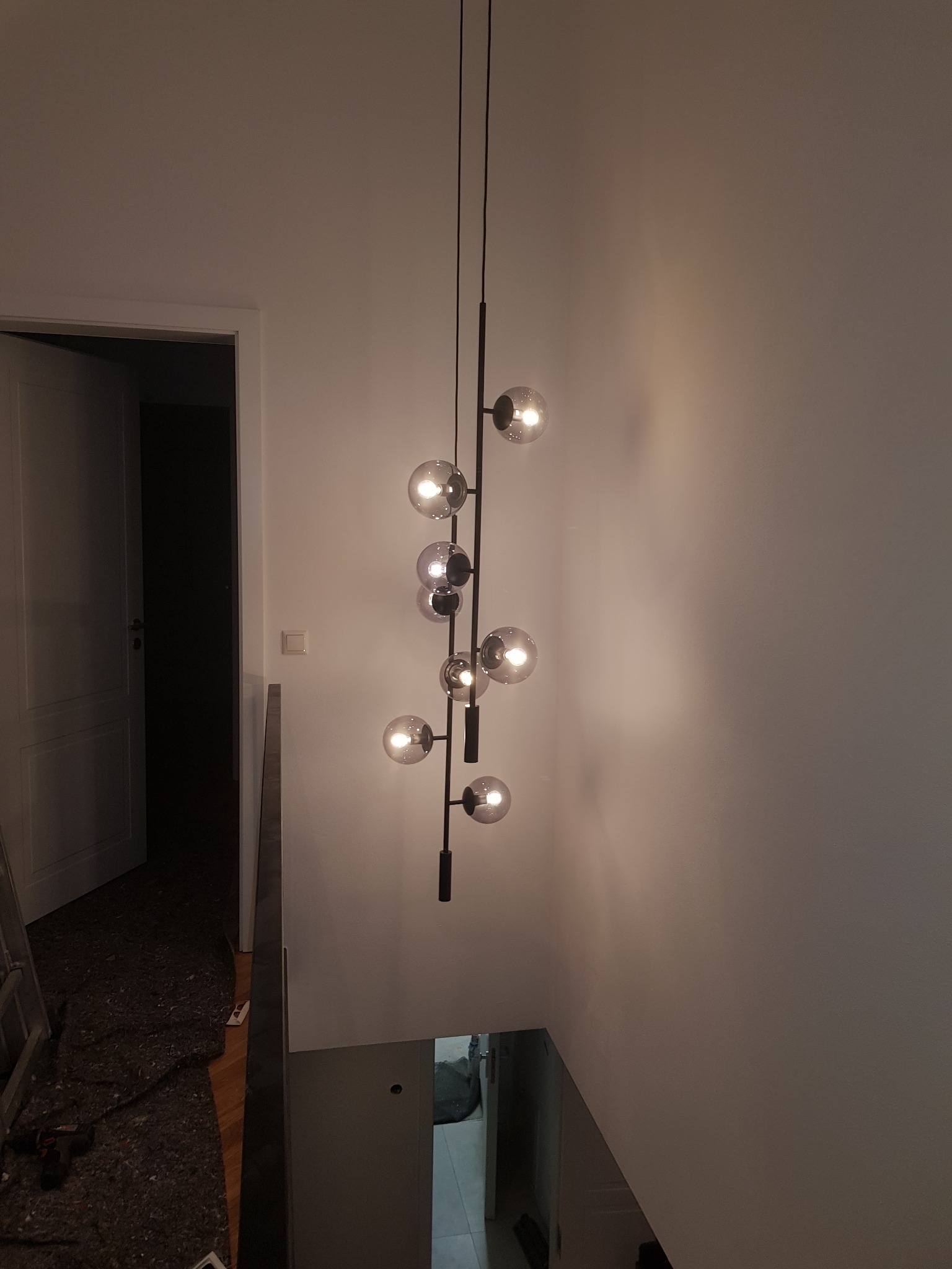 Nowoczesna lampa wisząca z kulistymi kloszami na czarnym stelażu, oświetlająca klatkę schodową w trakcie wykańczania wnętrza, widoczny fragment schodów i otwarte drzwi do pomieszczenia.
