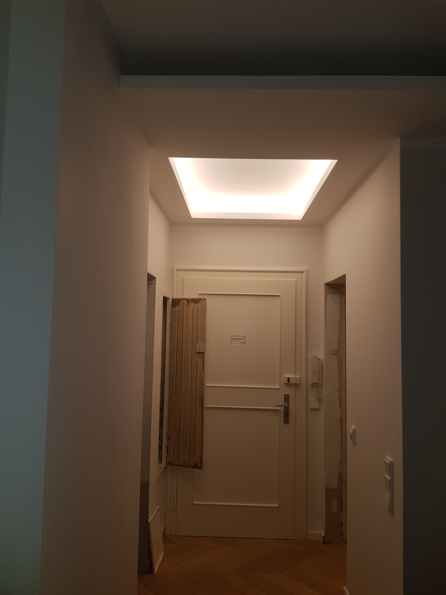 Korytarz w trakcie remontu z widocznym sufitem podwieszanym z oświetleniem LED, częściowo zasłoniętym oknem i przygotowanym otworem drzwiowym.