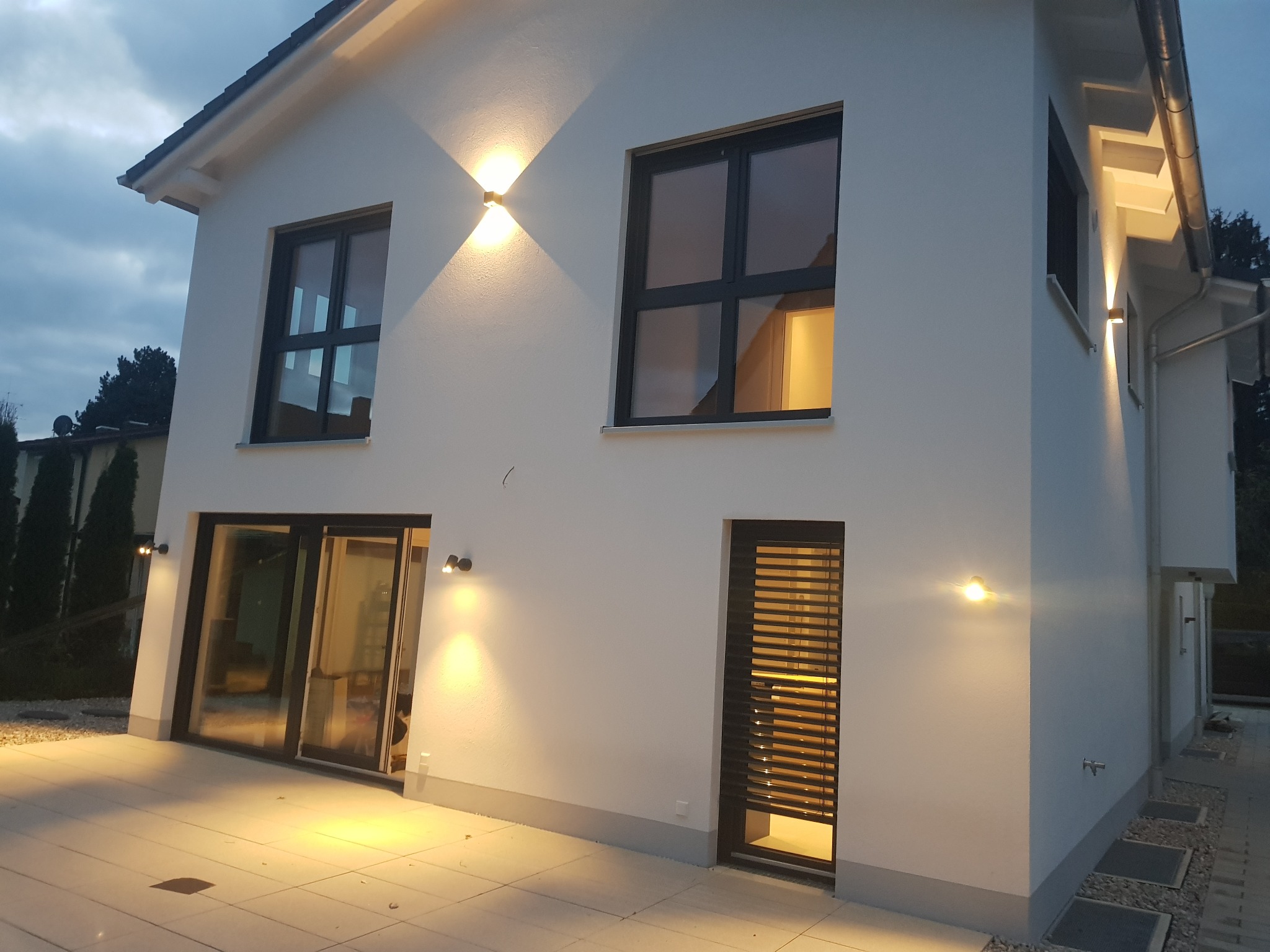 Elewacja domu z oświetleniem zewnętrznym, widoczne okna z czarnymi ramami, drzwi z żaluzjami, płytki na tarasie i małe lampy elewacyjne