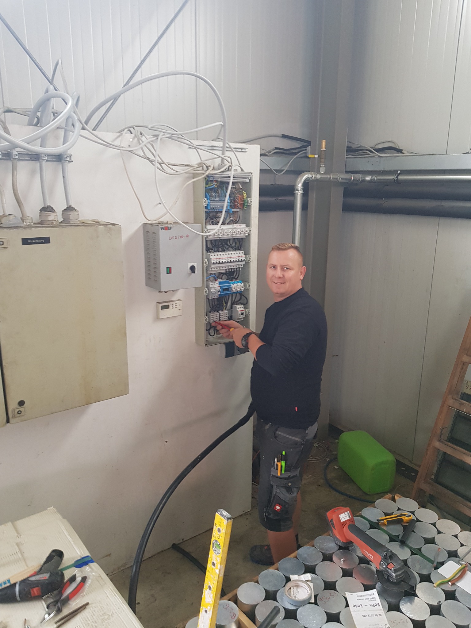 Elektryk uśmiecha się, pracując przy otwartej skrzynce z bezpiecznikami w przemysłowym wnętrzu; widoczne narzędzia, kable i elementy instalacji elektrycznej.