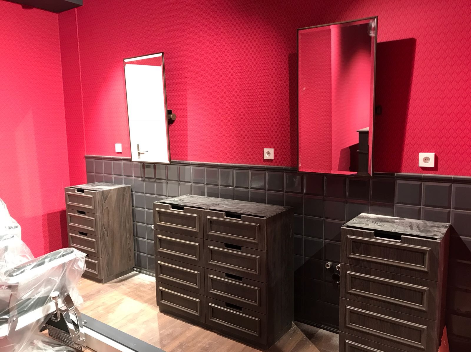 Łazienka w trakcie wykańczania: czerwona ściana z wzorem, szare płytki, trzy drewniane szafki z szufladami bez blatów, lustra i gniazdka elektryczne, widoczne rury instalacji wodnej.