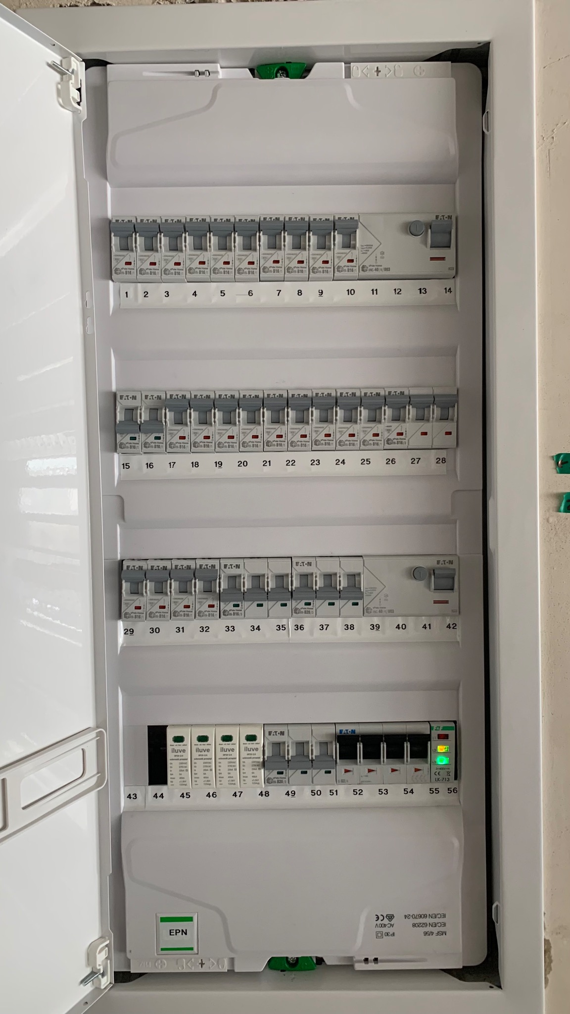 Biała skrzynka rozdzielcza z bezpiecznikami i wyłącznikami, numery od 1 do 56, niektóre w pozycji włączonej, widoczne detale producentów Eaton i Iluve.