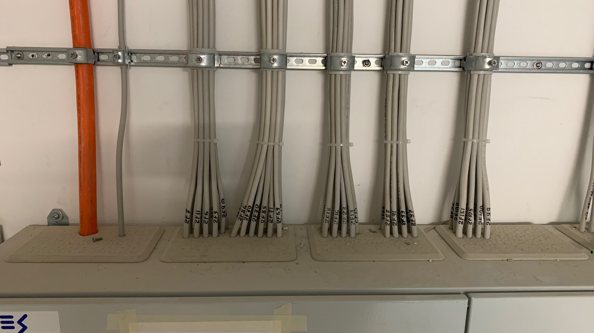 Szereg kabli oznaczonych numerami, uporządkowanych w wiązki, przymocowanych do metalowej szyny. Pomarańczowa rura w tle. Estetyczna instalacja.