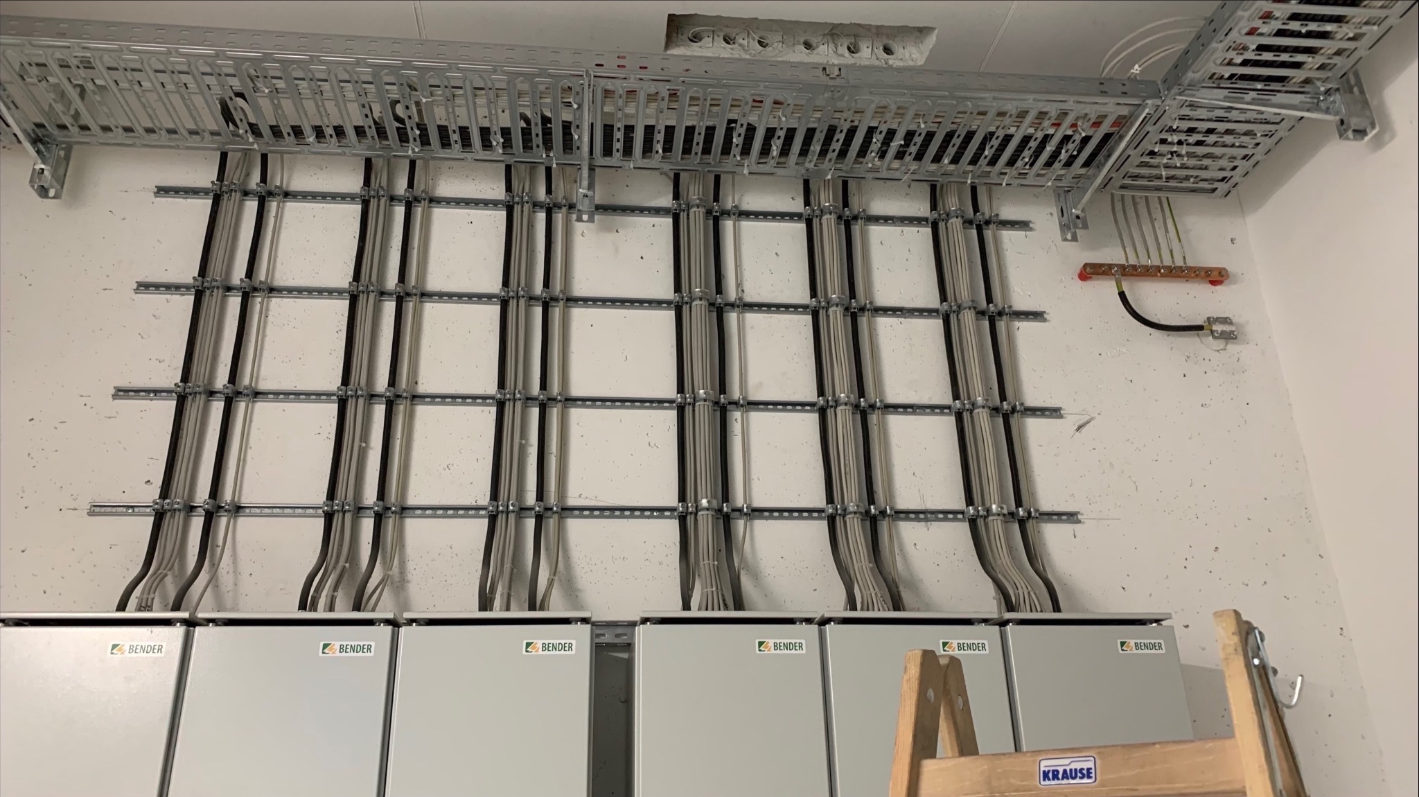 Szara ściana z zamontowanymi kablami w metalowych korytkach i szynach. Pod spodem zamontowane szare skrzynki elektryczne z logo BENDER. Fragment drabiny w prawym dolnym rogu.
