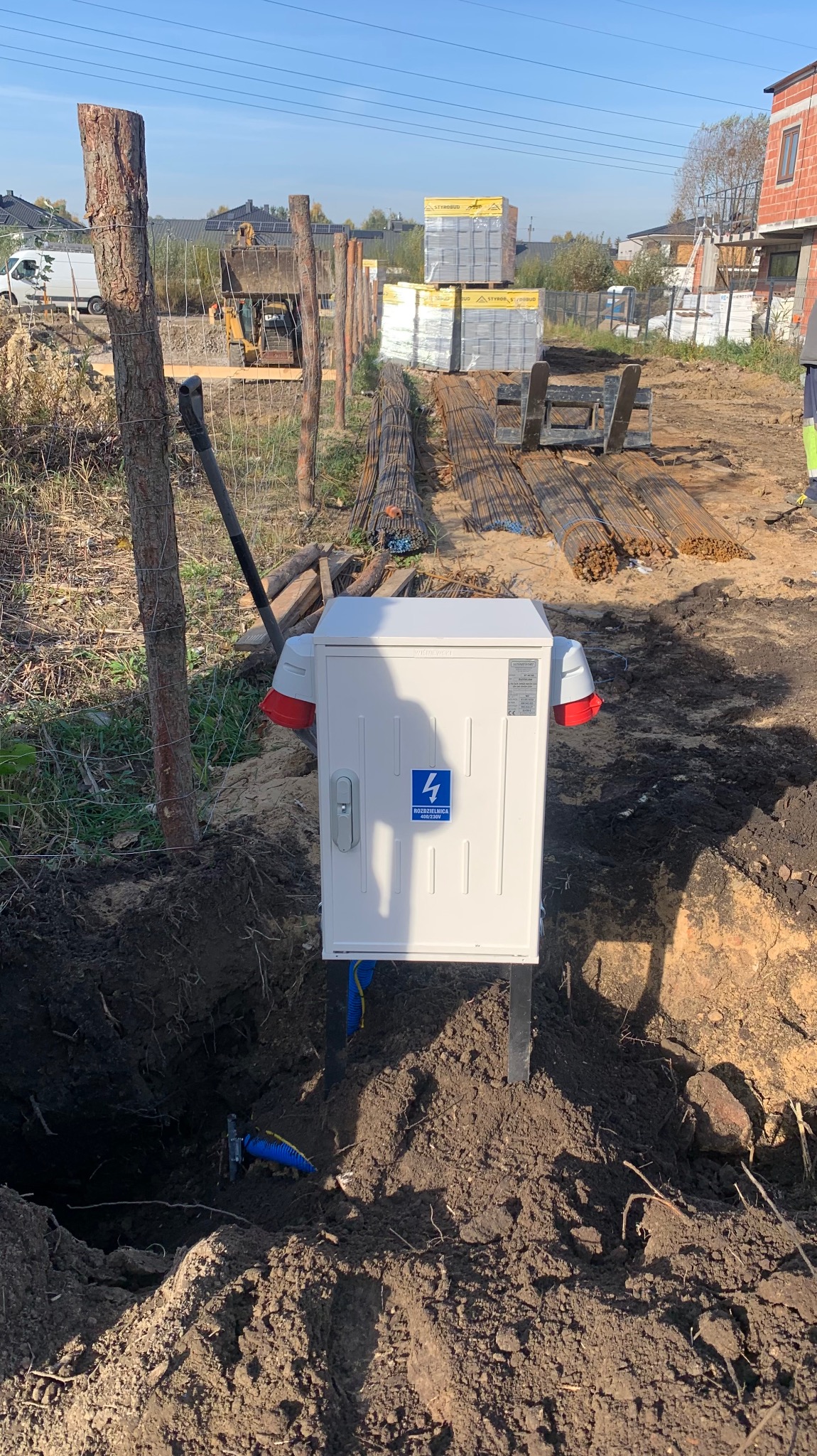 Biała skrzynka rozdzielcza z niebieską naklejką umieszczona w wykopie na placu budowy, widoczne przewody i elementy konstrukcyjne w tle.