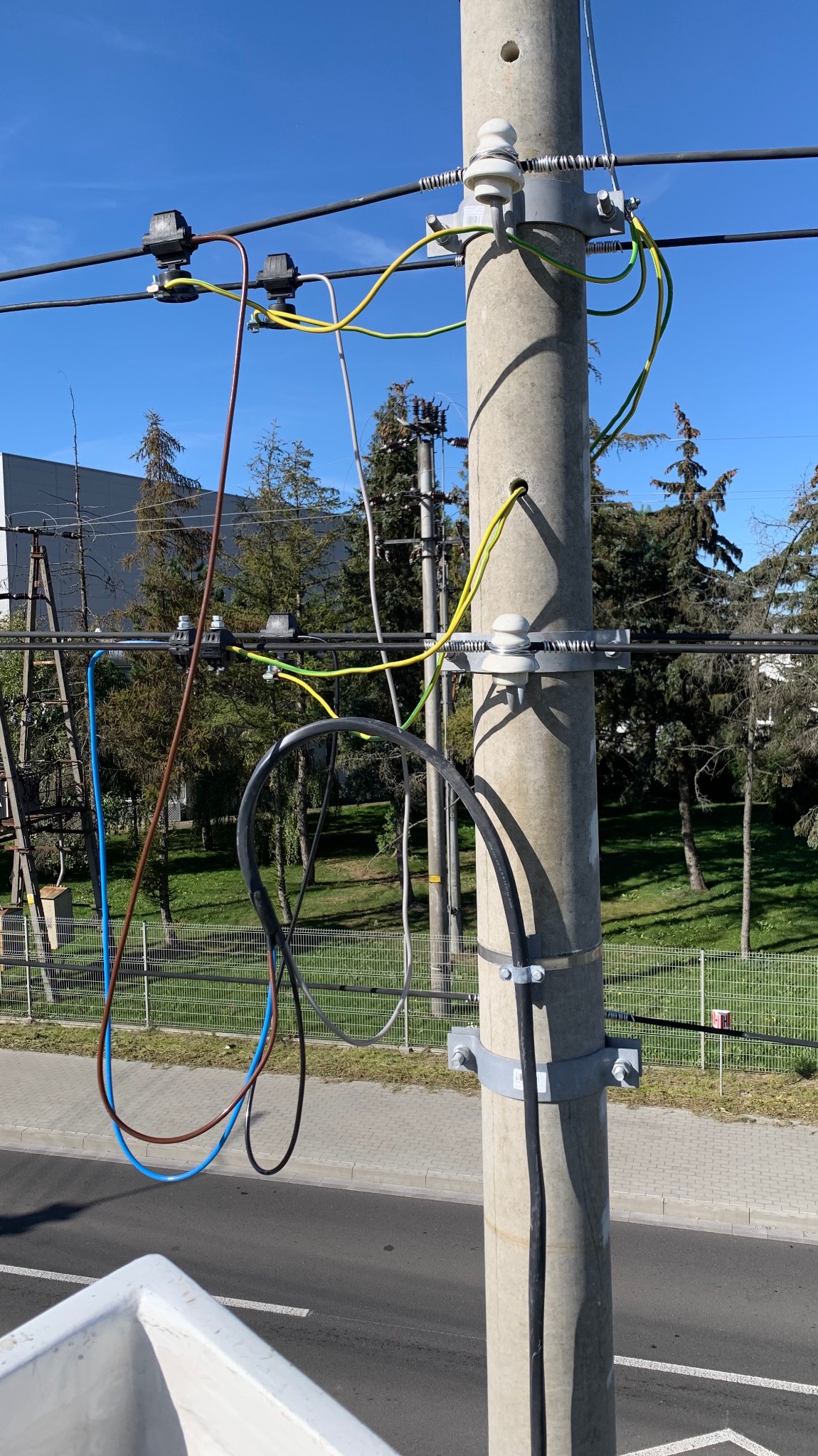 Betonowy słup energetyczny z zamontowanymi izolatorami i podłączonymi przewodami elektrycznymi, widok z dołu na tle błękitnego nieba i zieleni.