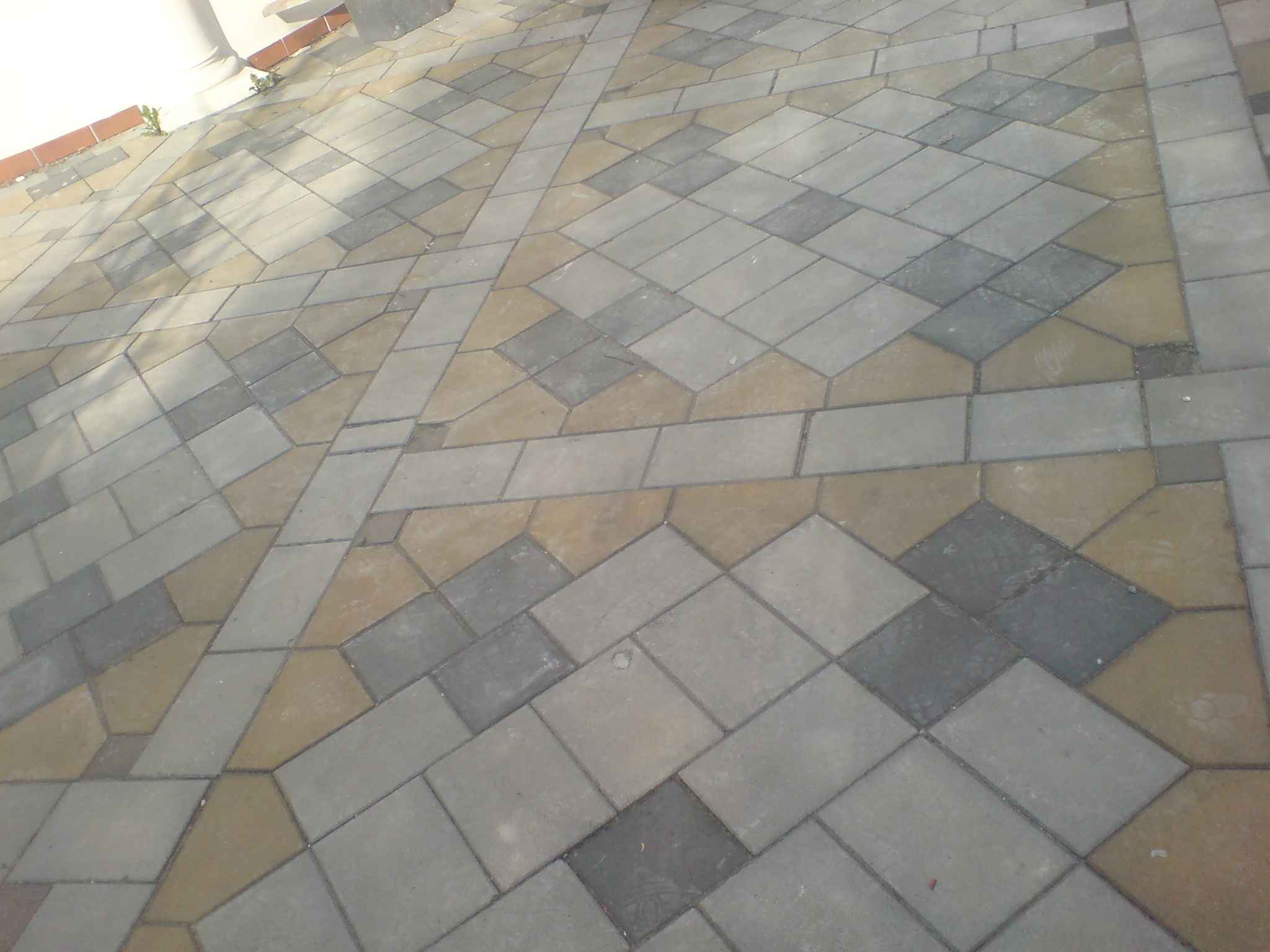 Ułożona nawierzchnia z kostki brukowej w odcieniach szarości i beżu, tworząca wzór geometryczny z elementami rombów i prostokątów, widok z góry pod kątem.