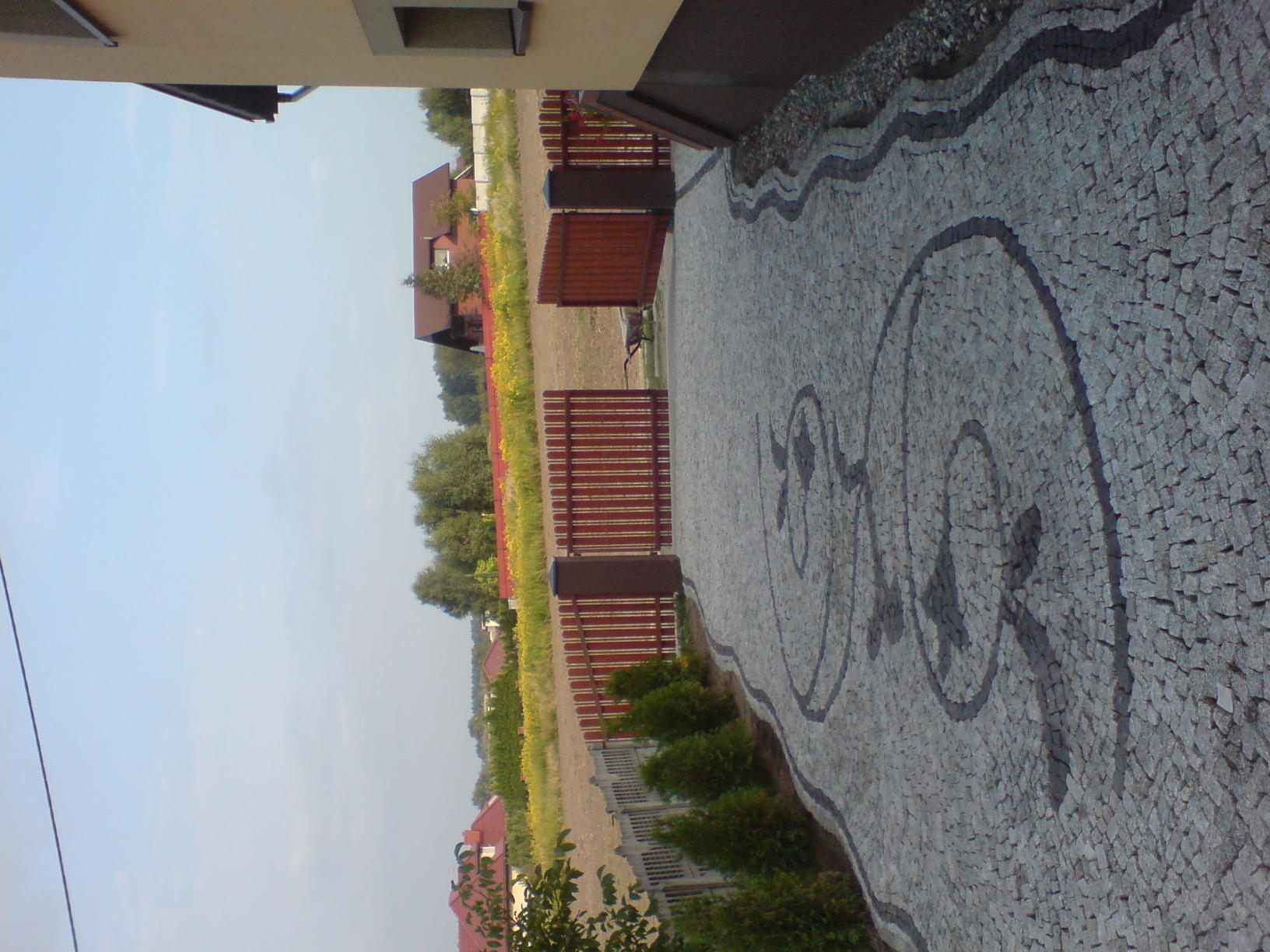 Ułożona z jasnoszarych kostek granitowych ścieżka z dekoracyjnym wzorem z ciemniejszych kostek, obok drewnianego ogrodzenia i niskich krzewów.