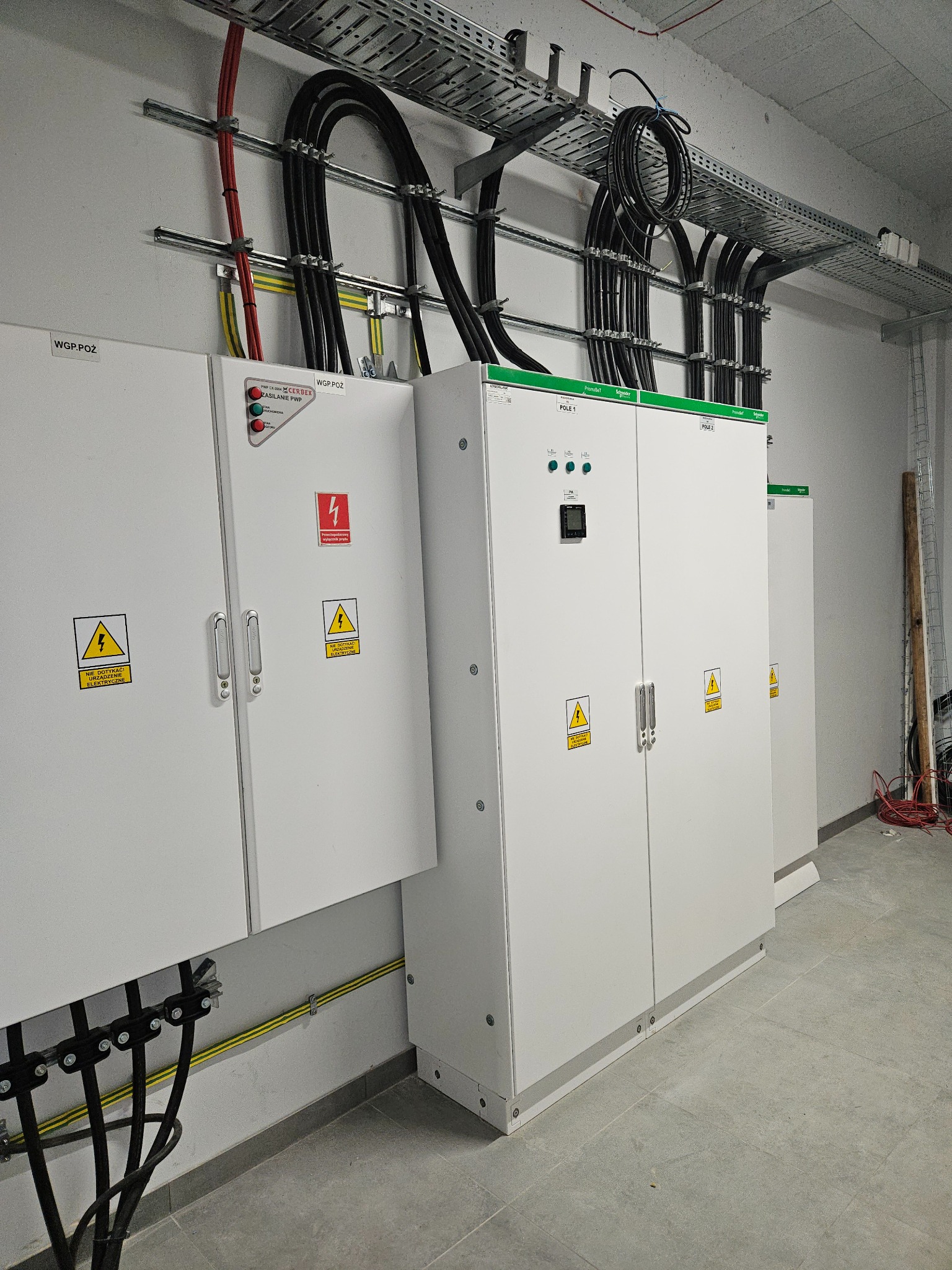 Rozdzielnice elektryczne w białych obudowach z ostrzeżeniami, kable prowadzone w korytach kablowych pod sufitem. Widoczne przewody w kolorach: czarnym, czerwonym i żółto-zielonym.