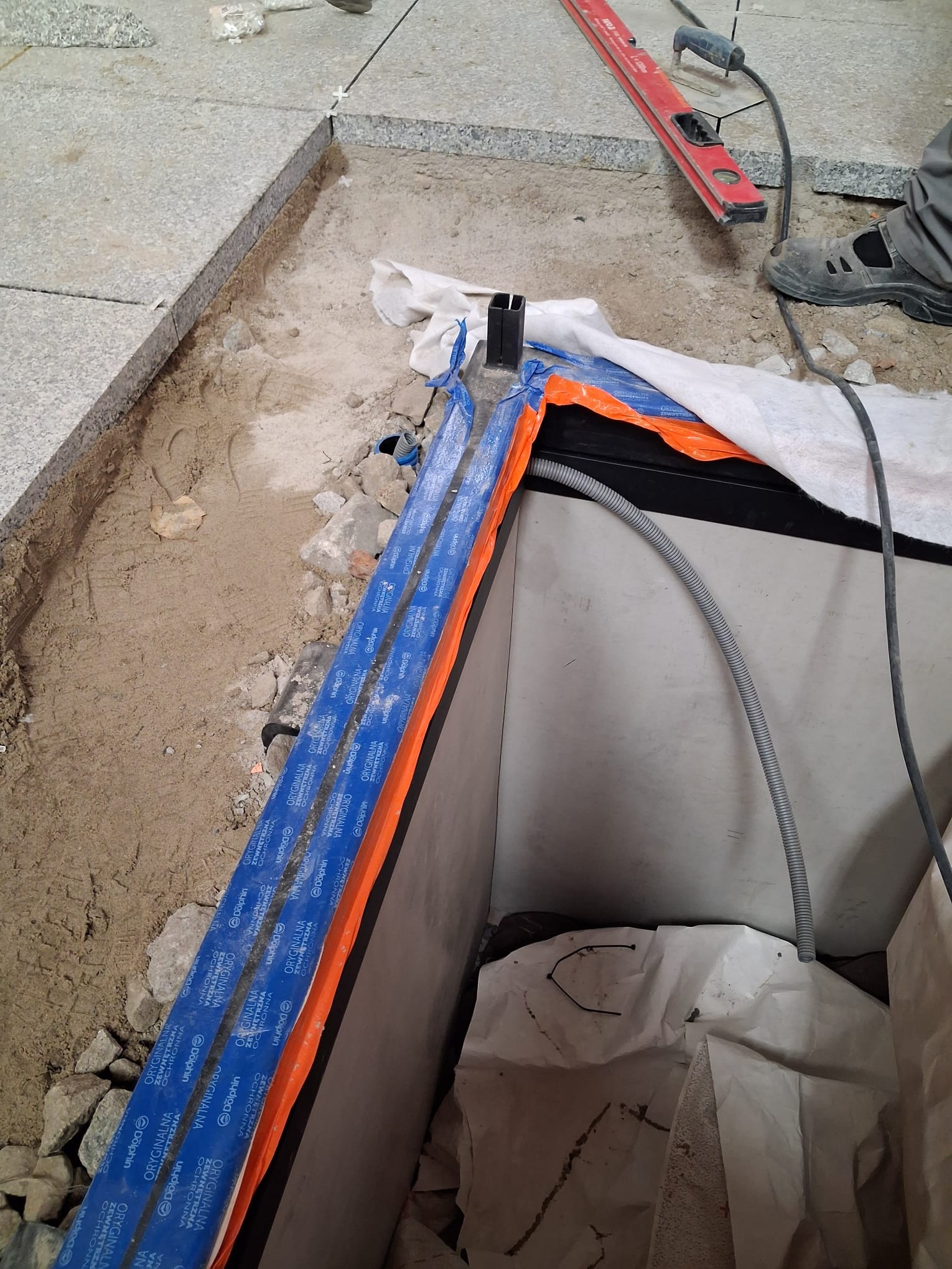 Przygotowanie do montażu oświetlenia w podłodze: kable w peszlach, poziomica, fragment ułożonej posadzki. Prace elektryczne w trakcie realizacji.