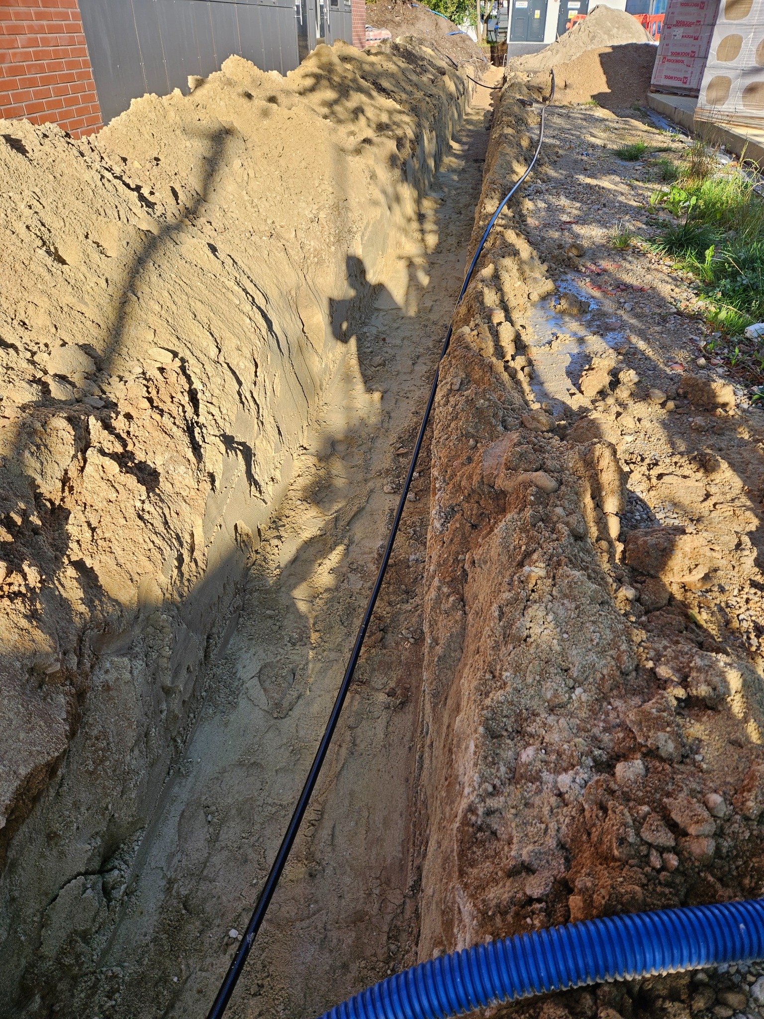 Wykop w ziemi z ułożonym kablem w peszlu, prawdopodobnie pod przyłącze. Widoczny cień osoby. Prace ziemne w trakcie realizacji.